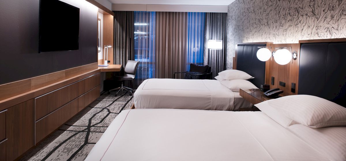 A modern hotel room with two double beds, a wall-mounted TV, a desk with a chair, and stylish decor, including patterned wallpaper and carpet.