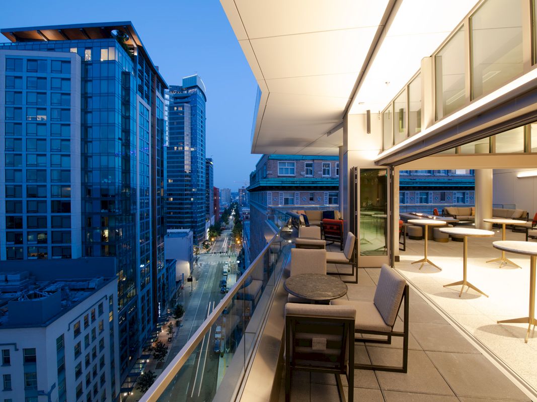 A modern cityscape at dusk featuring a high-rise balcony with seating, tables, and city lights, overlooking a street lined with tall buildings.
