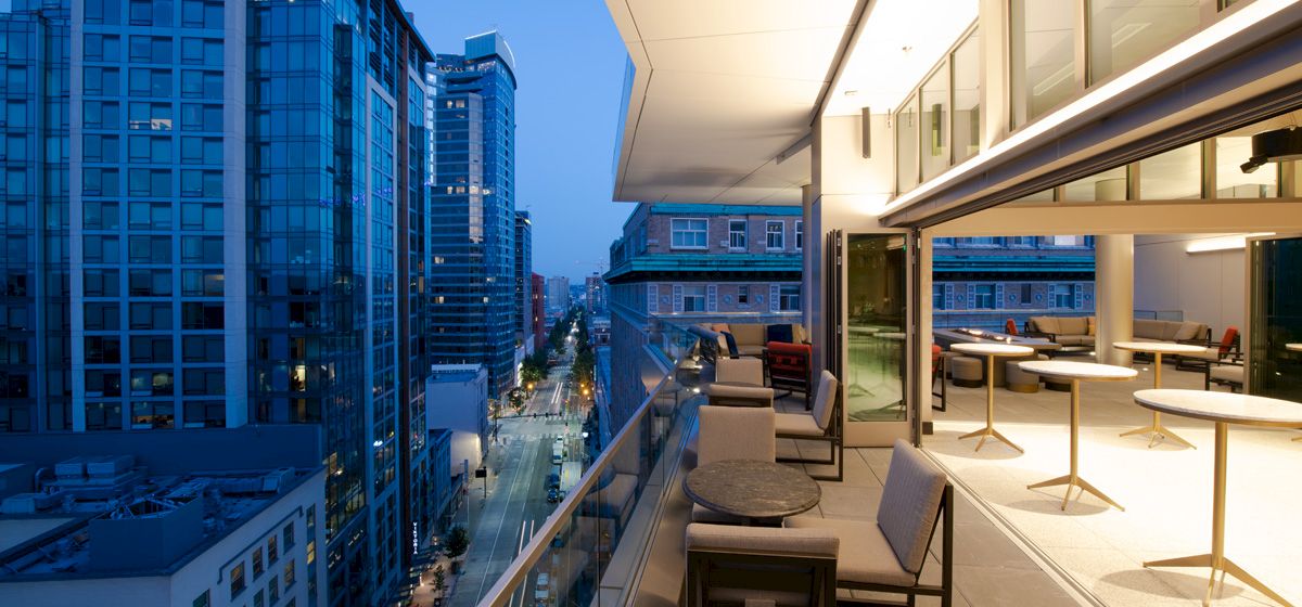 A modern cityscape at dusk featuring a high-rise balcony with seating, tables, and city lights, overlooking a street lined with tall buildings.
