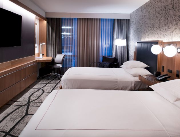 This image shows a modern hotel room with two beds, a TV, a desk with a chair, and stylish decor and lighting.