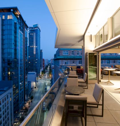 A modern balcony with furniture overlooks a cityscape featuring tall buildings at dusk. The interior is lit, and the scene feels tranquil.