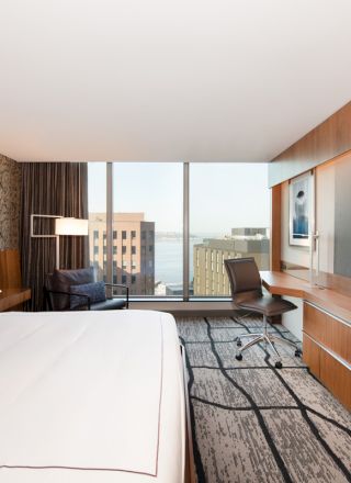 Modern hotel room with a large bed, desk, chair, flat-screen TV, cityscape view through floor-to-ceiling windows, and stylish decor, ending the sentence.