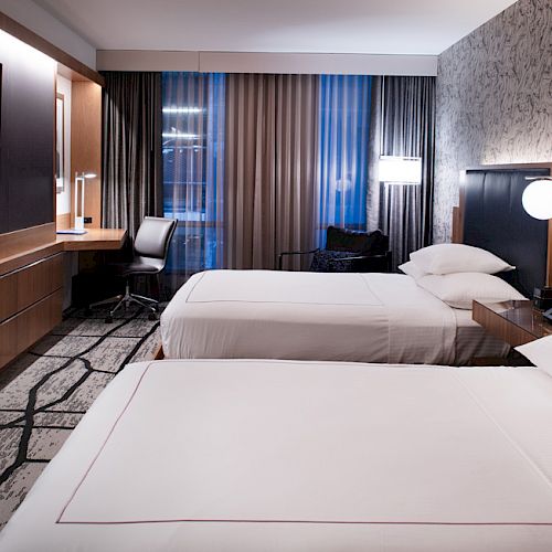 A hotel room featuring two neatly made beds, a desk with a chair, modern lighting, a flat-screen TV on the wall, and large windows with curtains.