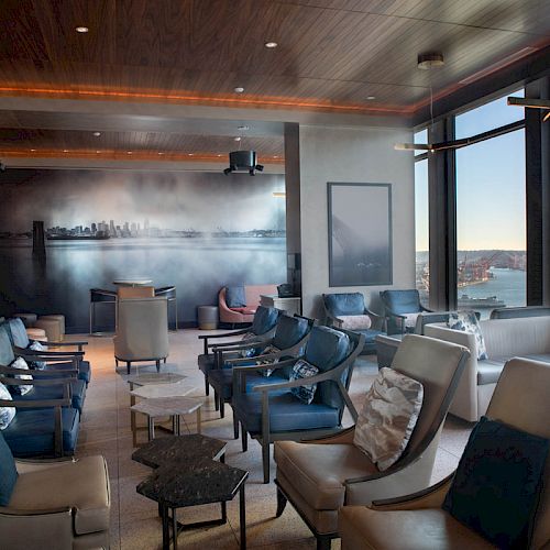 A modern lounge with large windows offering a scenic water view, featuring comfortable chairs, low tables, and a wall mural of a city skyline.