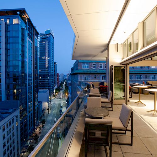 A modern balcony with seating and tables overlooks a cityscape at dusk, featuring tall glass buildings and a lit street below.
