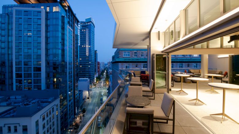 A modern rooftop patio with seating and tables overlooks a cityscape with tall buildings at dusk. The patio has glass railings and indoor lighting.