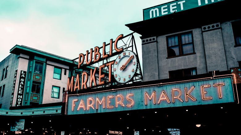 A public market sign with a clock and a neon "FARMERS MARKET" sign, surrounded by buildings, seen in an outdoor urban setting.