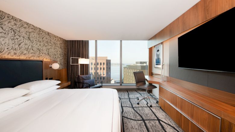 A modern hotel room with a king-sized bed, desk, chair, large TV, and a window offering a city and waterfront view.