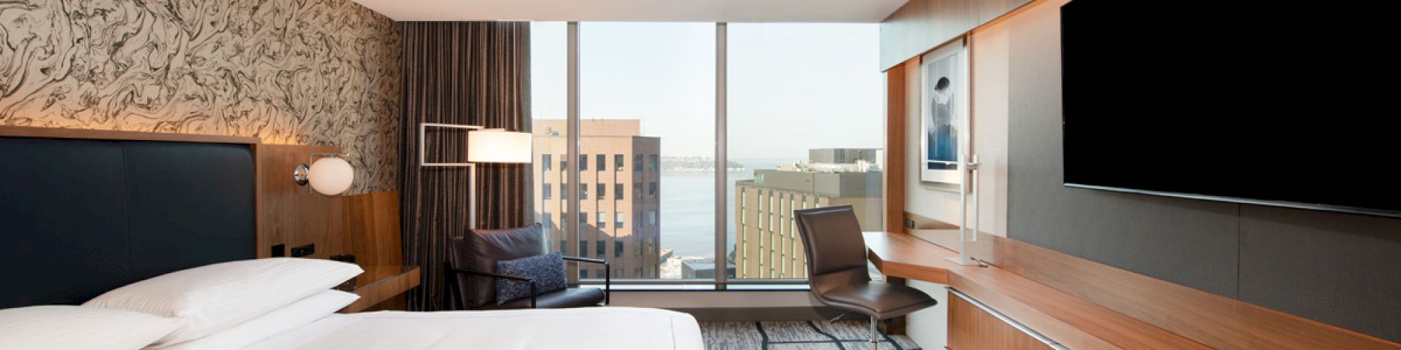 A modern hotel room features a large bed, wall art, a desk with a chair, a flat-screen TV, and a window with a city view.