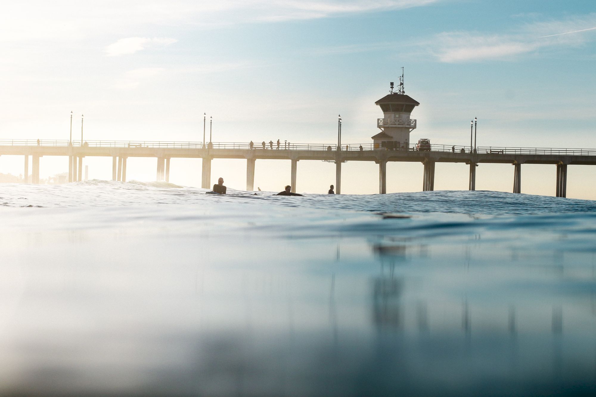 A serene beach scene with calm waters, a few people swimming, and a long pier with a tower extending into the ocean under a clear blue sky.